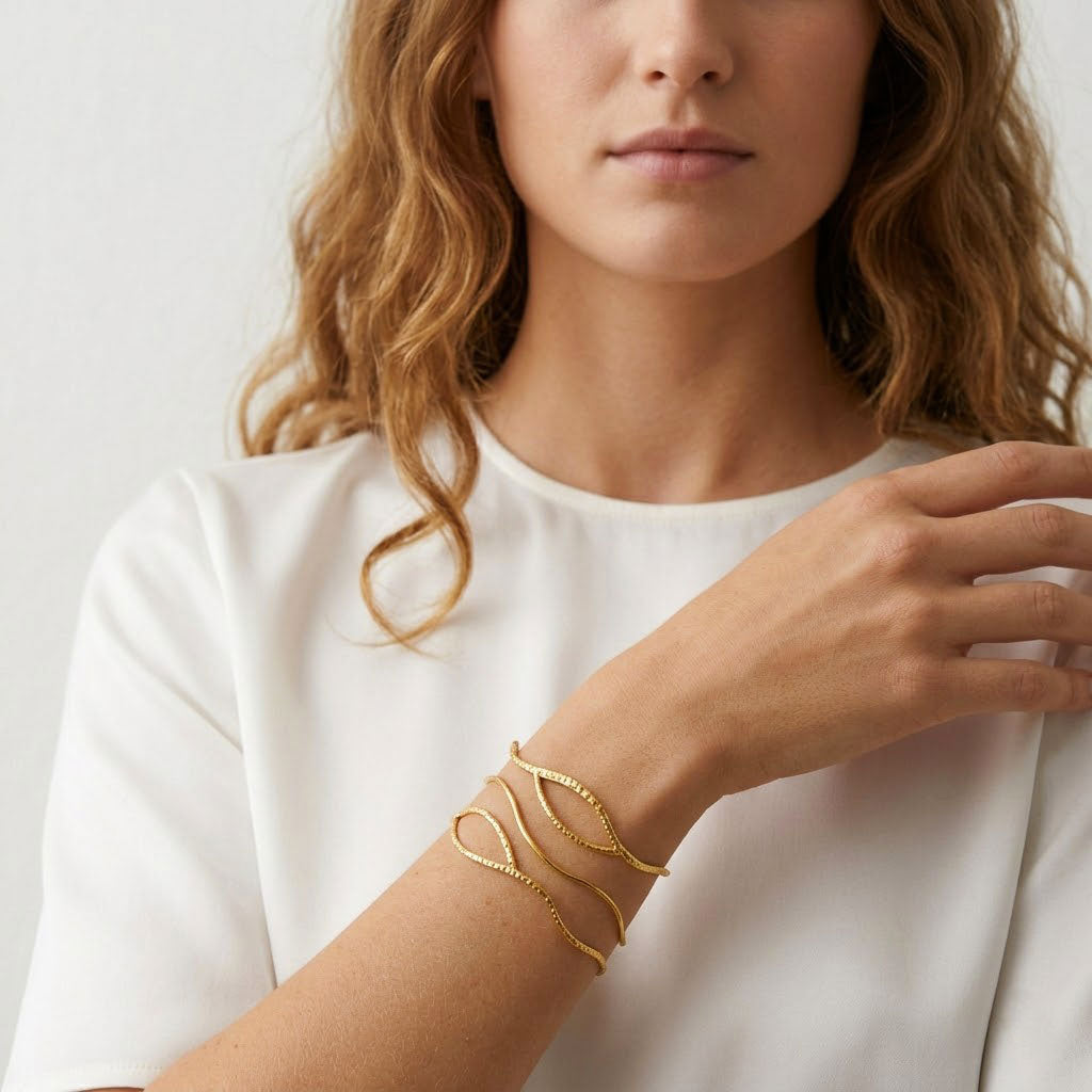 Brazalete abierto dorado con líneas onduladas y textura diamantada puesto en muñeca con camiseta blanca. Pulsera Anuska de Algo de Olga.