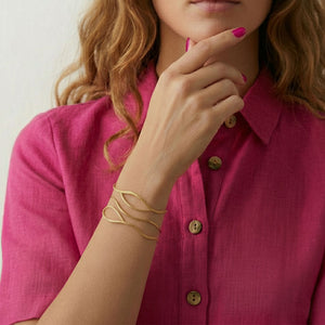 Pulsera tipo Brazalete dorado ondulado Anuska de acero inoxidable, la pulsera está puesta en la muñeca de la modelo con una camisa fucsia. Joyería Algo de Olga.