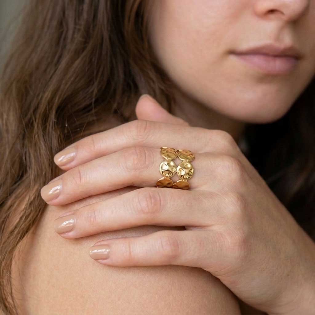 Mano con anillo Elena de acero dorado martillado con monedas superpuestas y uñas en tono nude.