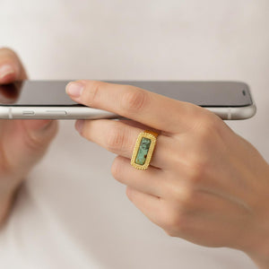 Mano sujetando un móvil con anillo de acero cepillado dorado y piedra turquesa